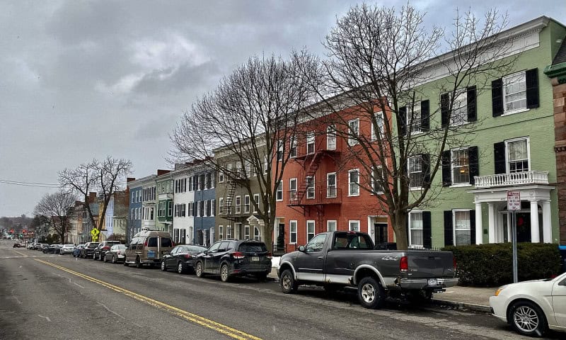 A row of painted brick houses along a residential street in Geneva, New York, with cars parked at the kerb
