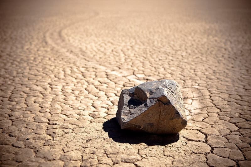 A pale tan and grey angular rock lit brightly on the cracked dry mud of Racetrack Playa, with a faint track running across the flat behind it