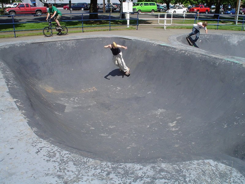 A skateboarder riding the curved concrete wall of a skatepark bowl, with a cyclist and parked cars behind