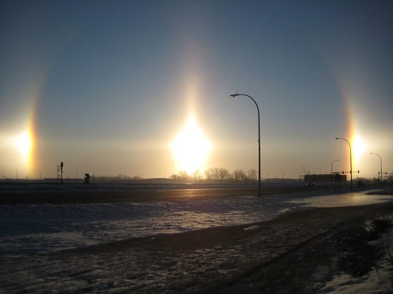 A low winter sun over a snow-covered road flanked by two bright sun dogs, with three tall vertical light pillars rising from the horizon