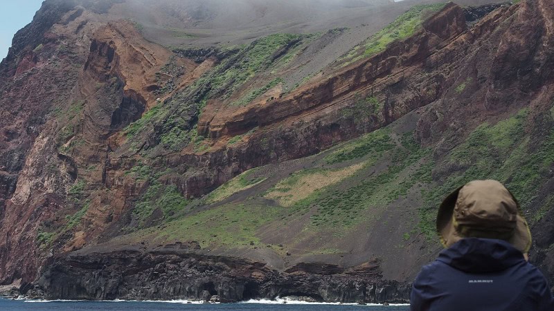 Layered brown and red volcanic cliffs of Torishima seen from a boat, with a hooded observer in the foreground