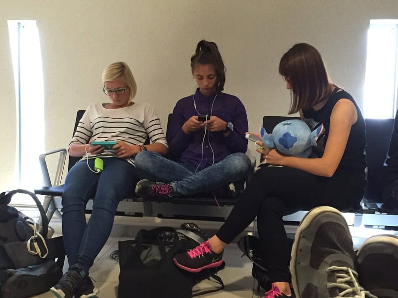 Three young people sitting side by side on airport seating, each looking down at a smartphone