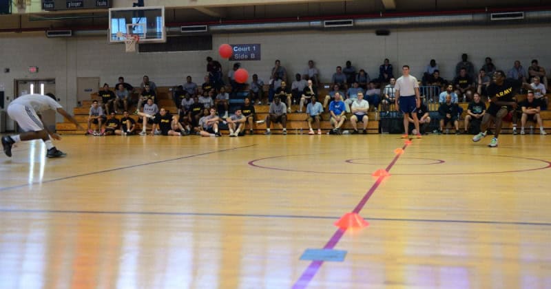 Players dodging a thrown ball on a gymnasium floor while a crowd watches from the bleachers