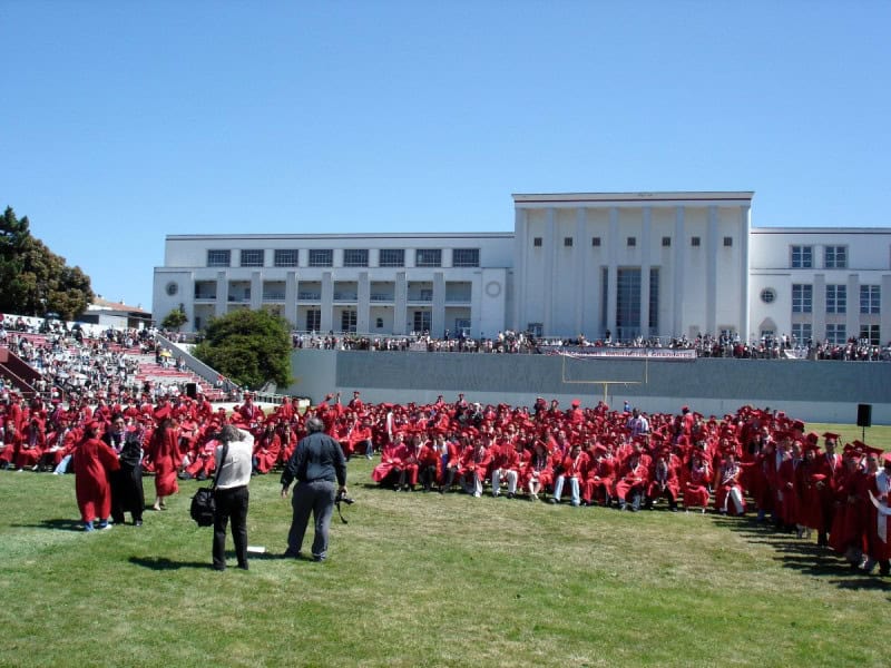 High school graduates in red gowns and caps seated in rows at an outdoor commencement ceremony, with spectators in the stands behind them