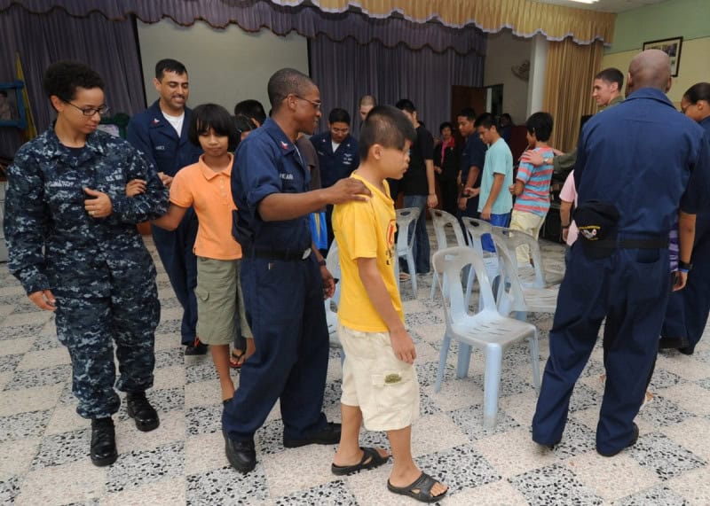 Students and sailors circling a row of chairs in a game of musical chairs in a school hall