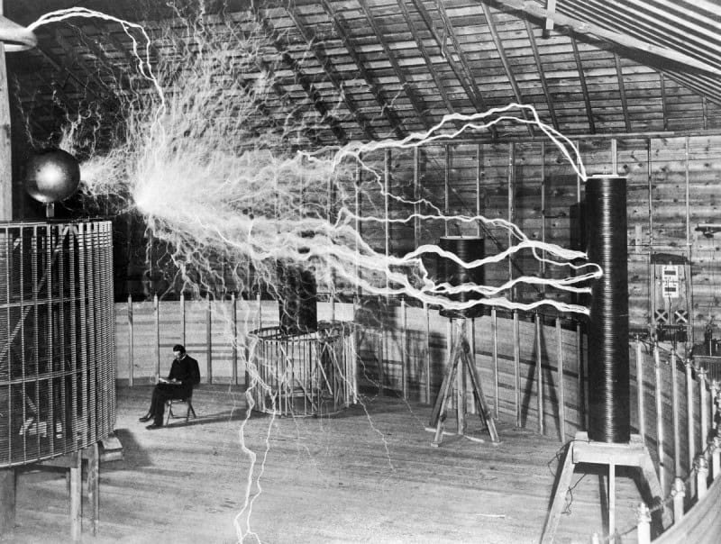 Nikola Tesla sitting and reading beside his magnifying transmitter in Colorado Springs in 1899 while long electrical discharges fill the room, a double-exposure publicity photograph