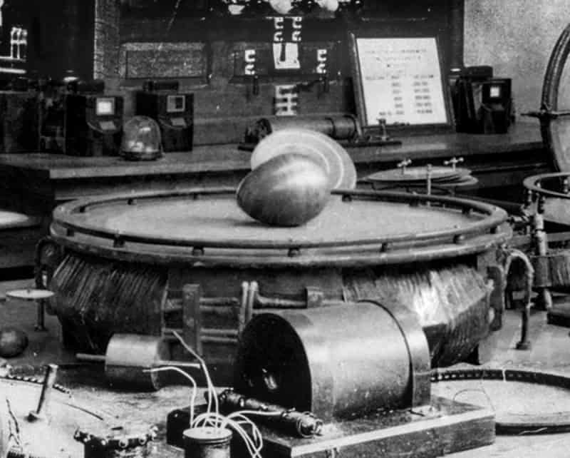 Photograph of Tesla's Egg of Columbus apparatus from 1893, a large copper egg resting on the round platform of a polyphase coil, with laboratory equipment behind