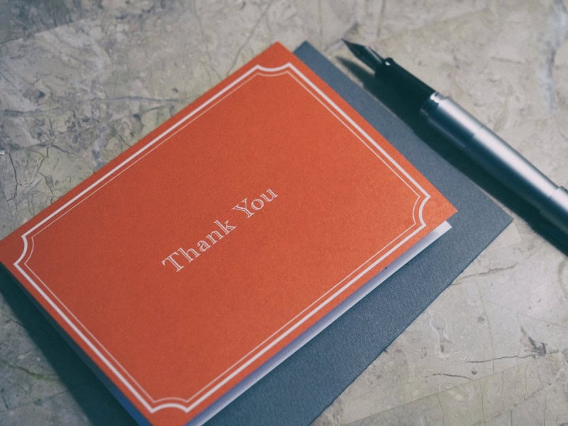 An orange thank-you card with a white border lying on a grey notebook next to a pen