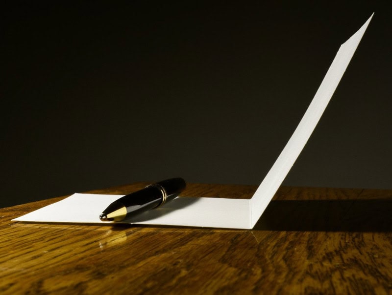 A folded white note card standing open on a wooden desk with a black fountain pen resting in front of it